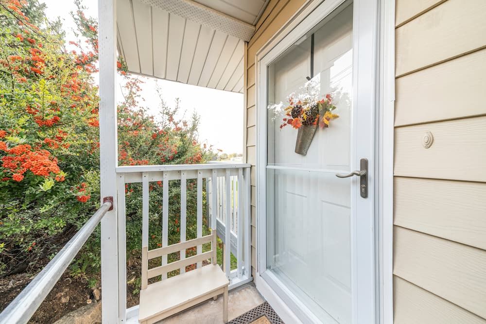 A small porch with a white railing, a beige exterior wall, a glass door decorated with a flower wreath, and a wooden bench beside red-berried bushes.