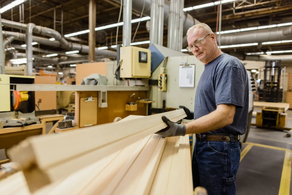 A man wearing gloves and safety glasses inspects wooden boards in a large industrial woodworking facility.
