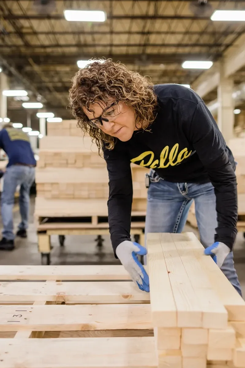 A person wearing safety glasses and gloves stacks wooden planks in a large indoor lumber facility. Another worker is visible in the background.