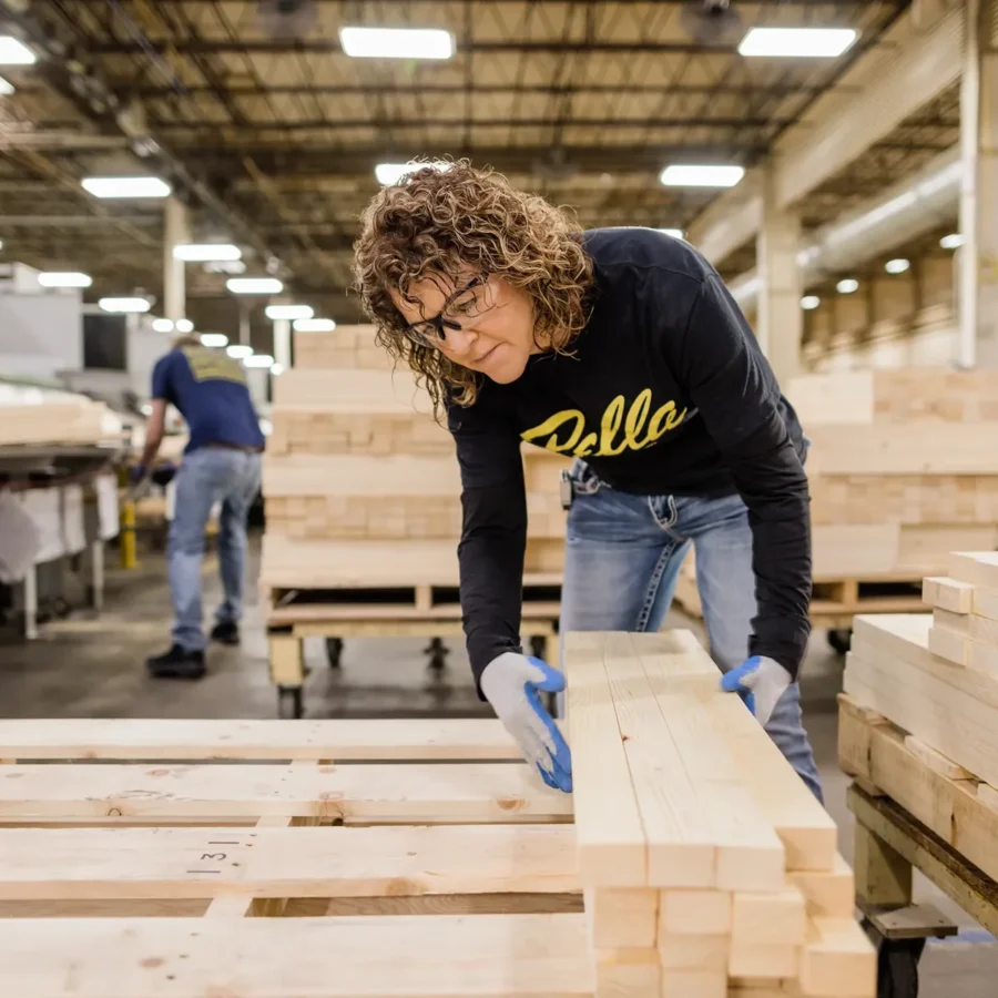 A person wearing safety glasses and gloves stacks wooden planks in a large indoor lumber facility. Another worker is visible in the background.
