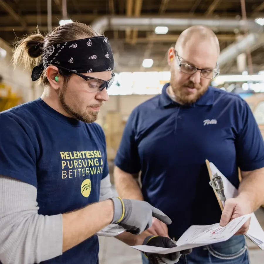Two men in a workshop review documents together; one is wearing safety glasses and gloves, the other holds a clipboard and pen.