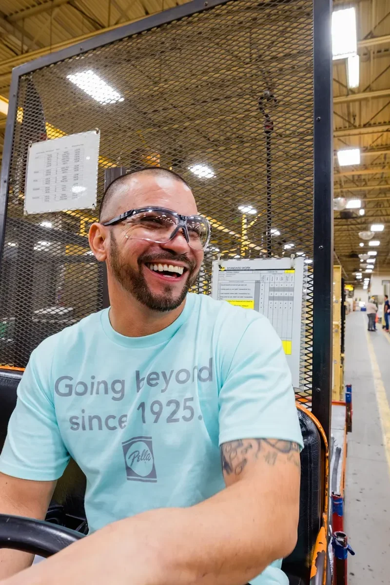 A man wearing safety glasses and a light blue T-shirt drives a vehicle inside a brightly lit factory or warehouse, smiling as he looks to the side.