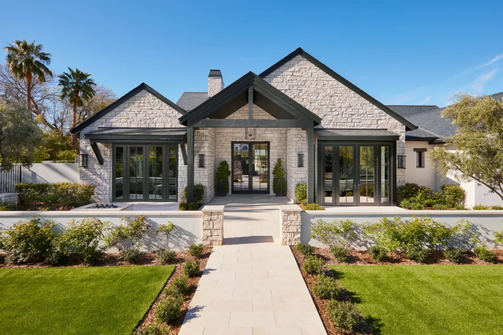 A modern stone house with black trim, large windows, a peaked entrance, and landscaped front yard under a clear blue sky.