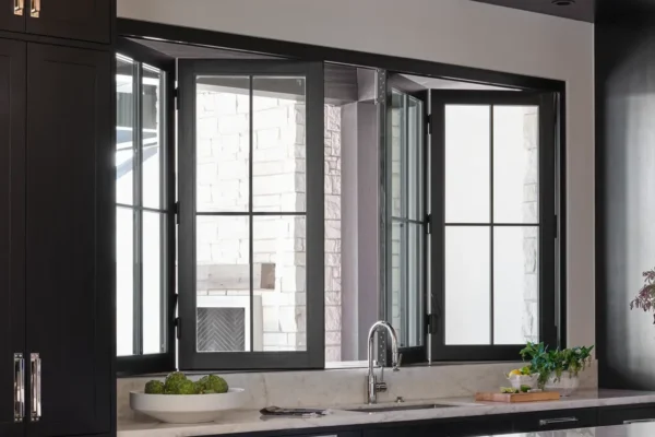 A kitchen sink with a modern faucet sits below large, open black-framed windows. A bowl of green apples and a cutting board with herbs are on the countertop.