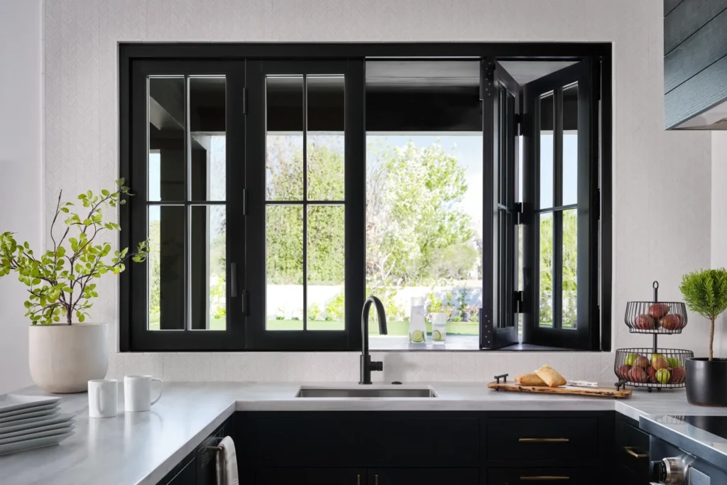 A modern kitchen with a black-framed window above the sink, open to a green outdoor view. The countertop holds dishes, a potted plant, and a tiered fruit stand.