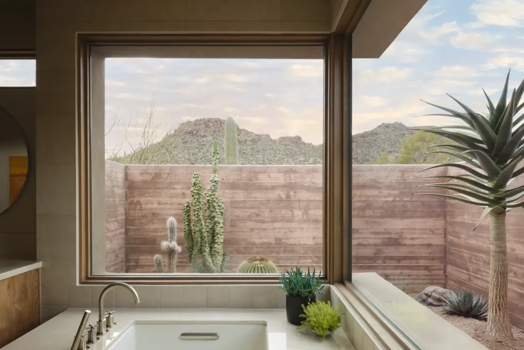 A modern bathroom sink with large windows overlooks a walled courtyard featuring various cacti and desert plants, with mountains and a partly cloudy sky in the background.