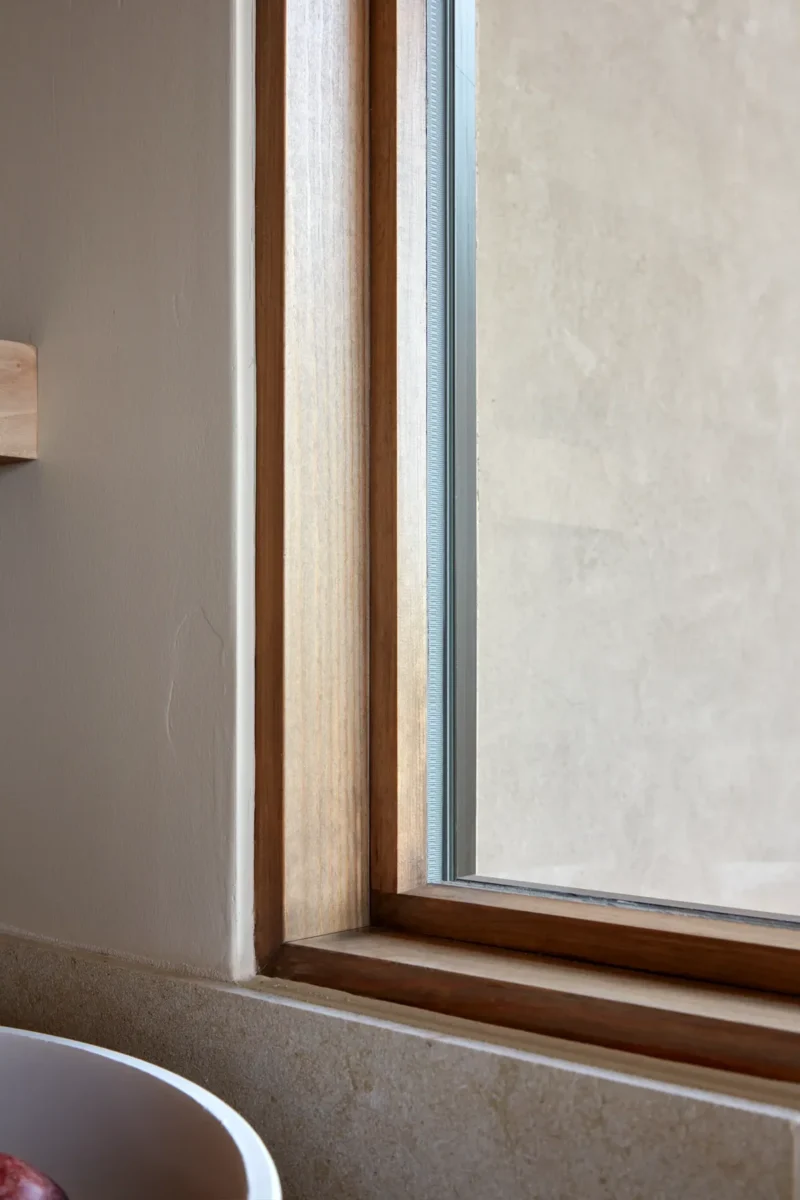 Close-up of a window with a wooden frame set in a beige wall, showing a partial view of a white countertop and an off-white exterior surface outside.