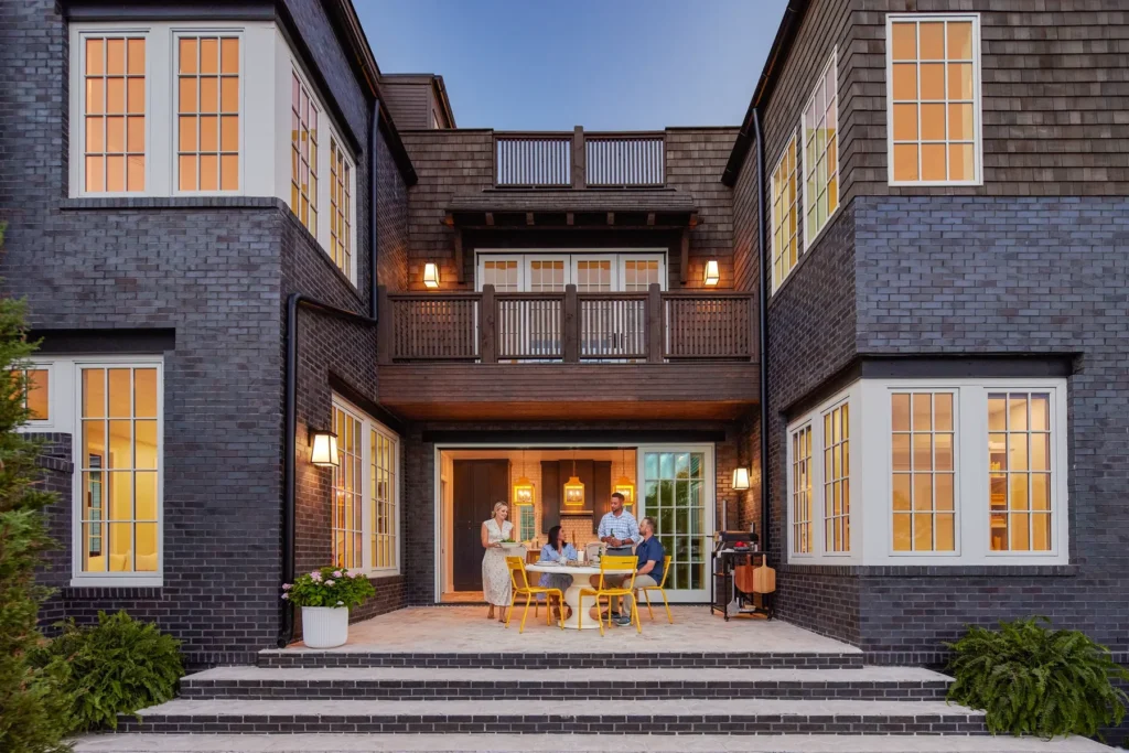 Three people sit at a yellow table on a brick patio, surrounded by a modern, dark-brick house with large windows and a balcony above.