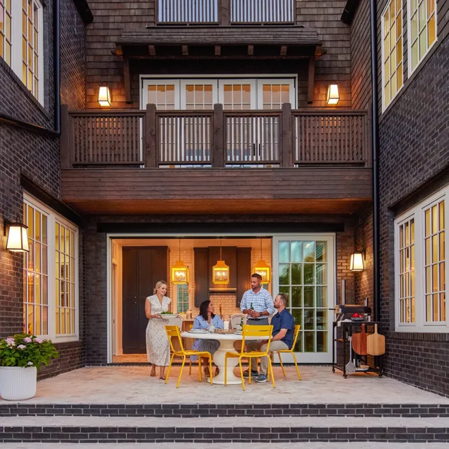 Four adults sit and stand around a table on a brick patio outside a house with large windows and yellow chairs, under evening lighting.