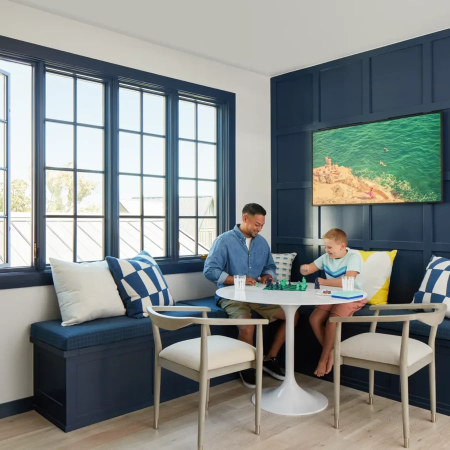 An adult and a child sit at a round table playing chess in a bright room with blue paneled walls and built-in bench seating.