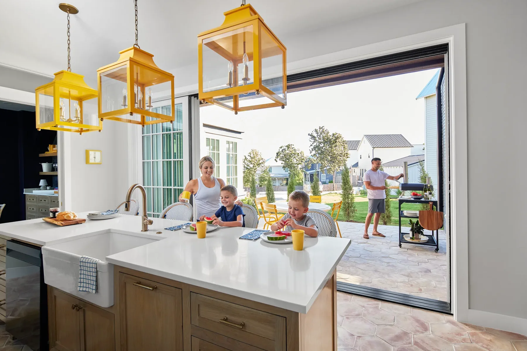 A family enjoys breakfast at a kitchen island while a man grills food outside on a patio with sliding glass doors open to the backyard.