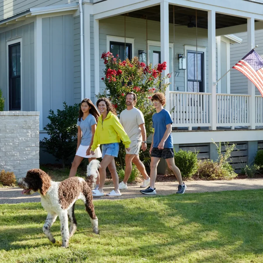 Four people walk together on a sidewalk in front of a house with an American flag, while a brown and white dog runs on the grass nearby.