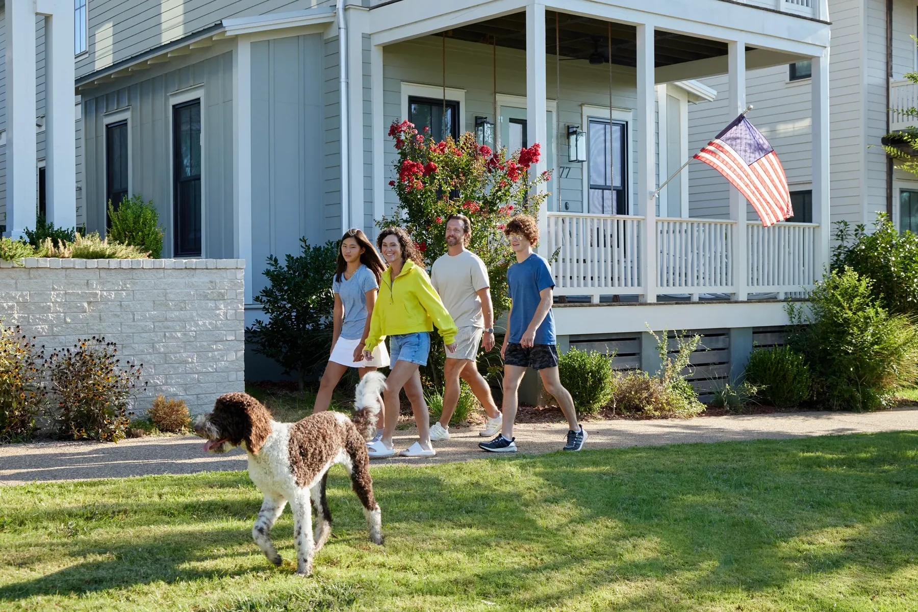 Four people walk together on a sidewalk in front of a house with an American flag, while a brown and white dog runs on the grass nearby.