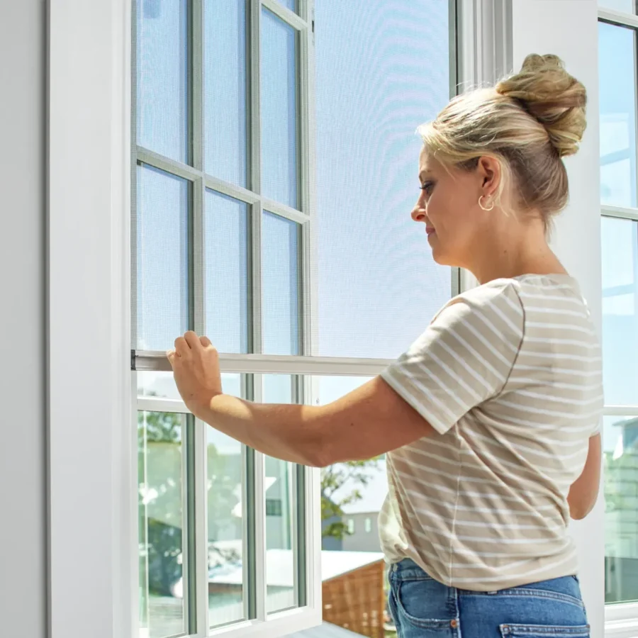 A woman installs or adjusts a window screen inside a bright room with large windows.