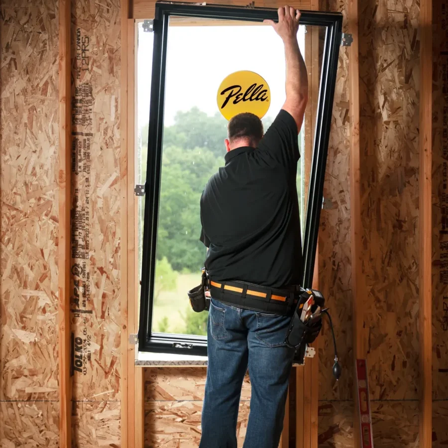 A worker installs a window frame in a house under construction, with exposed wooden studs and a tool belt around his waist.