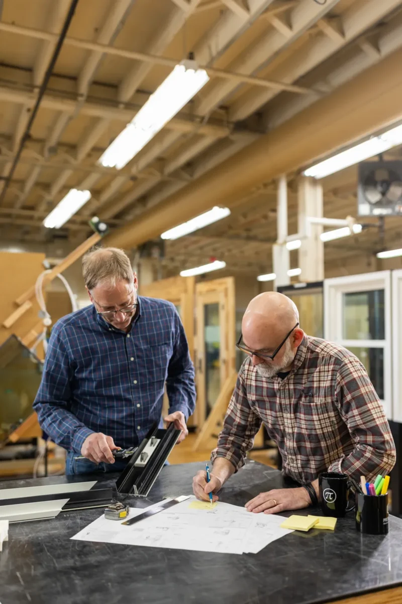 Two men in plaid shirts work together at a table, reviewing blueprints and using tools in a well-lit industrial workshop.