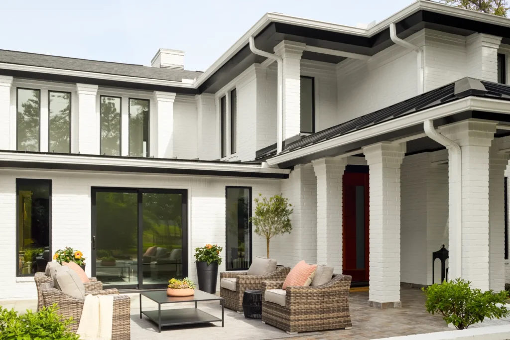 Modern white two-story house with large windows, a red front door, patio area with outdoor furniture, and potted plants.