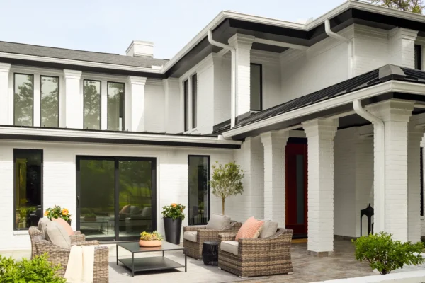 Modern white two-story house with large windows, a red front door, patio area with outdoor furniture, and potted plants.