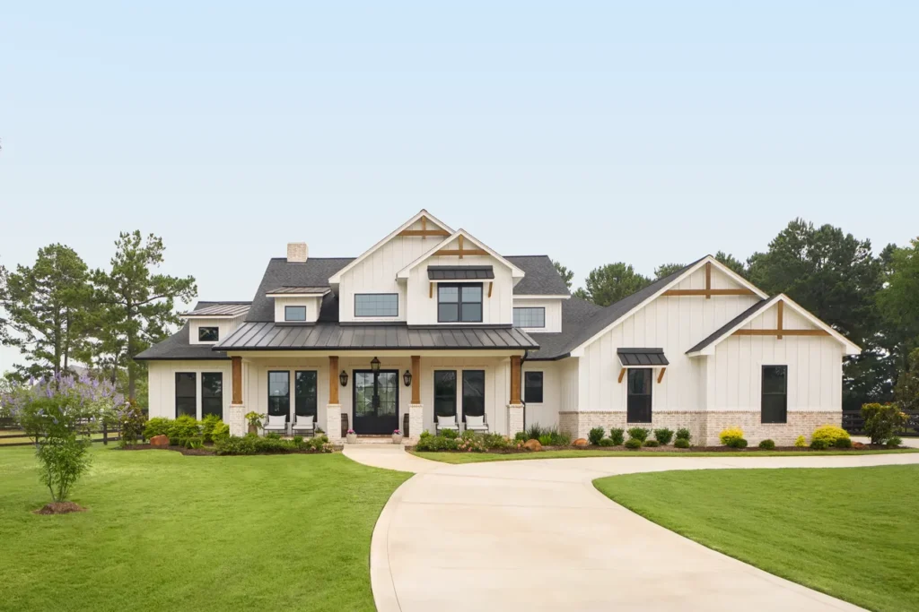 A modern farmhouse with white siding, black windows, a dark metal roof, and a landscaped yard, viewed from the front with a curved driveway.