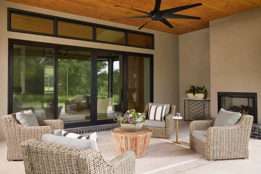 Covered patio with wicker chairs, a round wooden coffee table, a rug, potted plants, and a ceiling fan. Glass doors open to an indoor living area.