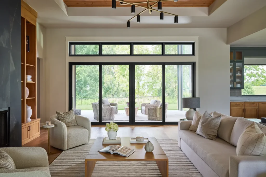 Modern living room with neutral sofas, a wooden coffee table, and large glass doors opening to a patio with chairs and a view of greenery outside.