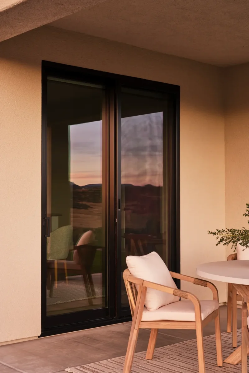 Glass sliding door reflecting an outdoor landscape at sunset, with a wooden chair and round table with a plant in the foreground.