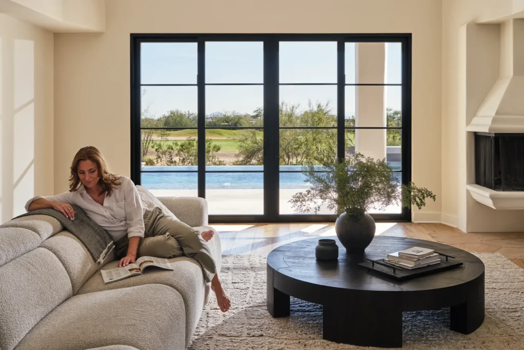 A woman relaxes on a light-colored sofa reading a book in a modern living room with large windows overlooking a pool and outdoor landscape.