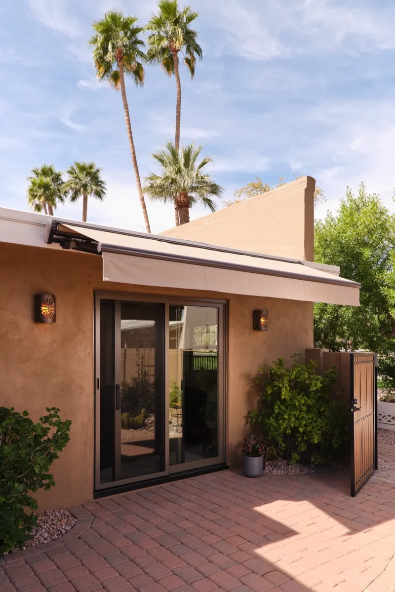 A sliding glass door with an extended awning opens to a brick patio, surrounded by green plants and palm trees under a partly cloudy sky.