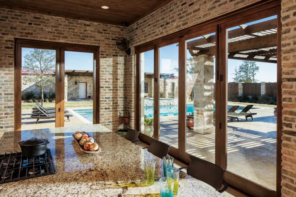 A kitchen with granite countertops and a gas stove overlooks a patio with lounge chairs and a swimming pool through large glass doors.