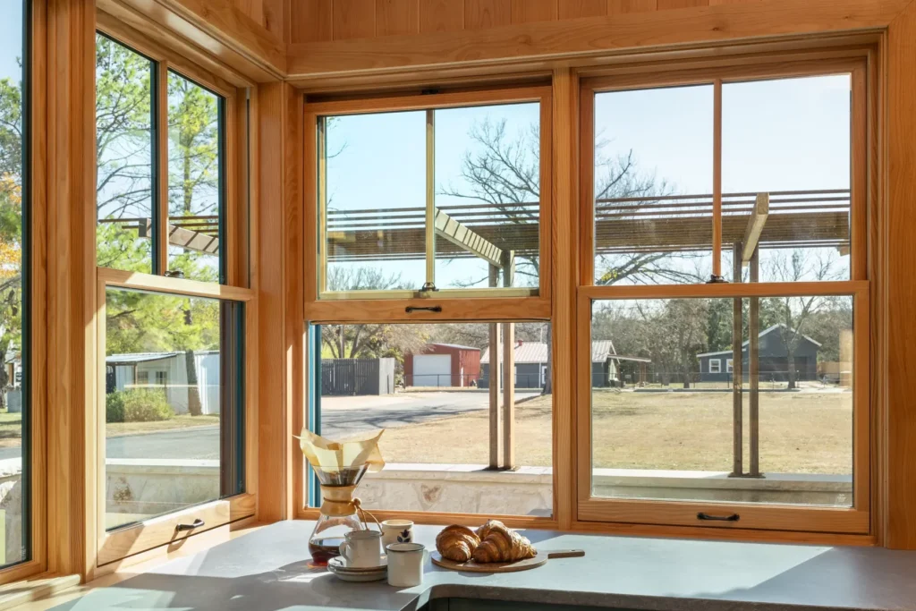 A corner kitchen window with light wood frames overlooks a yard; croissants, a coffee maker, and cups sit on the countertop in sunlight.