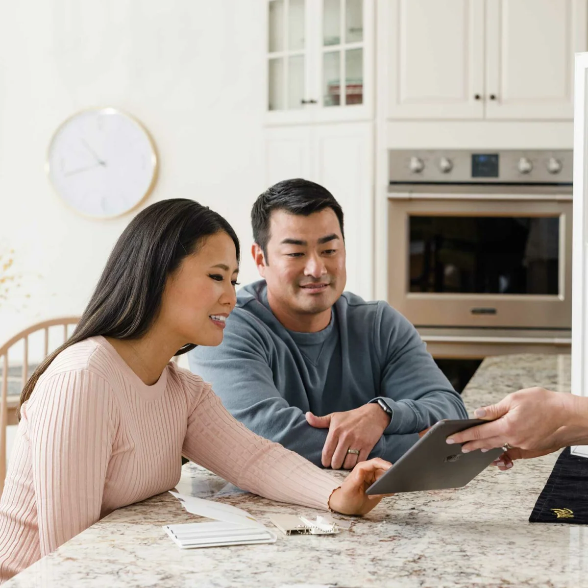 Two people sit at a kitchen counter, looking at a tablet device held by a third person. A notebook and pen are on the counter, possibly for taking notes about window replacement options, and a wall clock is visible in the background.