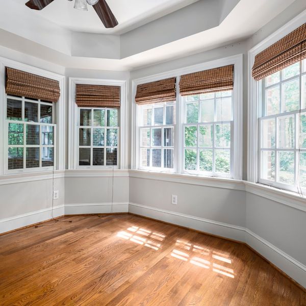 Empty room with hardwood floors, white walls, five large windows with woven shades, and sunlight streaming in from outside. Ceiling fan visible at the top.