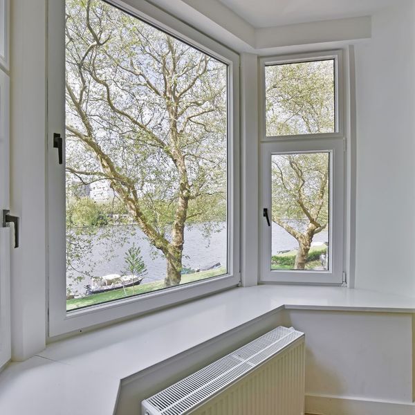 Large bay window with white frames overlooks a river and leafy trees. A white radiator is positioned below the window inside a light-colored room.