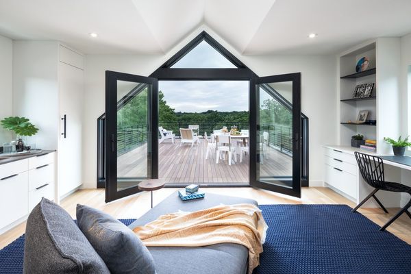 A modern bedroom opens to a spacious wooden deck with outdoor seating, overlooking green trees under a cloudy sky.