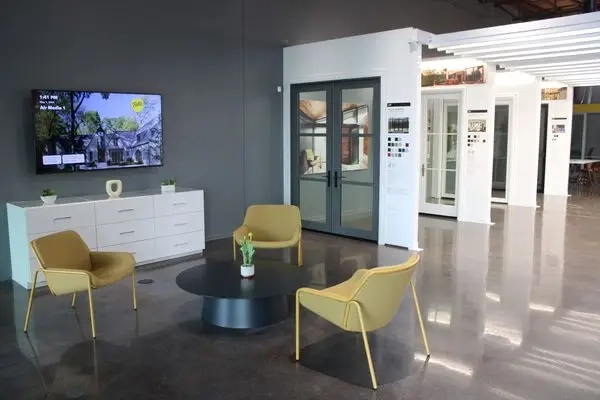 Modern showroom with glass doors, display sections, a TV on the wall, white cabinet, and four yellow chairs around a round black table on a polished concrete floor.