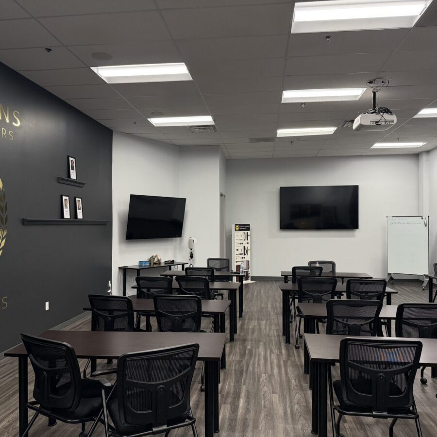 Modern classroom with rows of desks and chairs, large wall displays, whiteboards, and a “CHAMPIONS” feature wall on the left. The room is well-lit with ceiling lights.