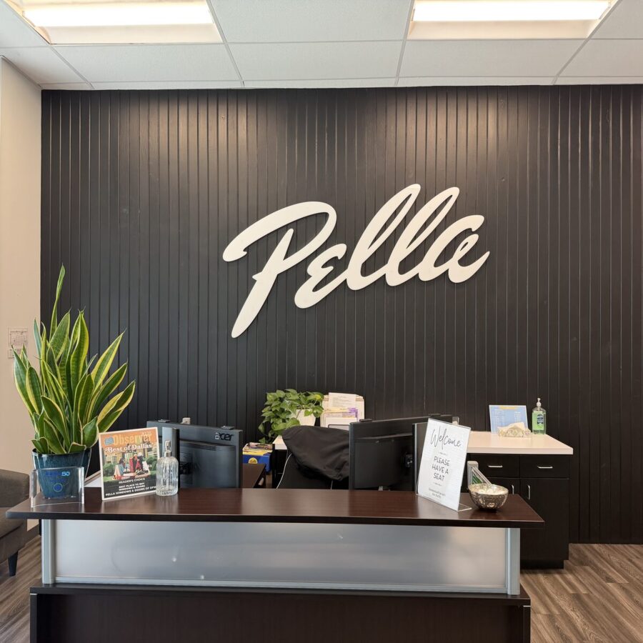 Reception desk in front of a black wall with the “Pella” logo, topped with office items, plants, a sign-in sheet, and hand sanitizer.