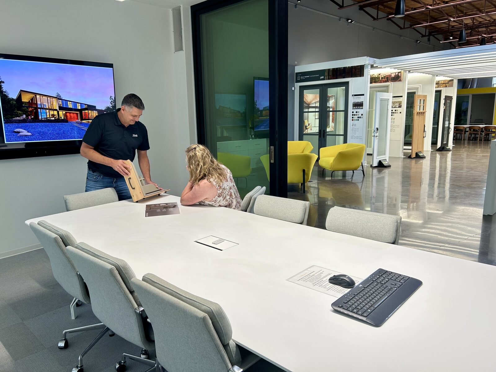 A man shows a wood sample to a seated woman in a modern office meeting room with a large table, chairs, and a TV screen displaying house images.