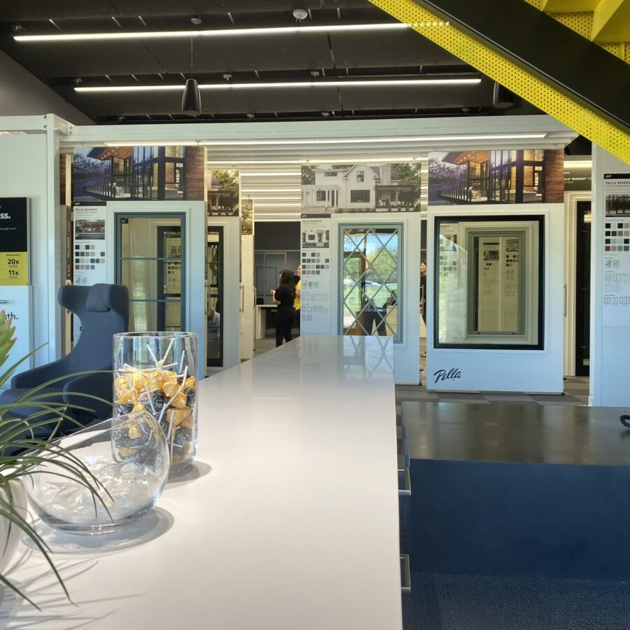 Modern showroom interior featuring window displays, a white table with a glass jar of snacks, a plant, and a yellow staircase under overhead lighting.