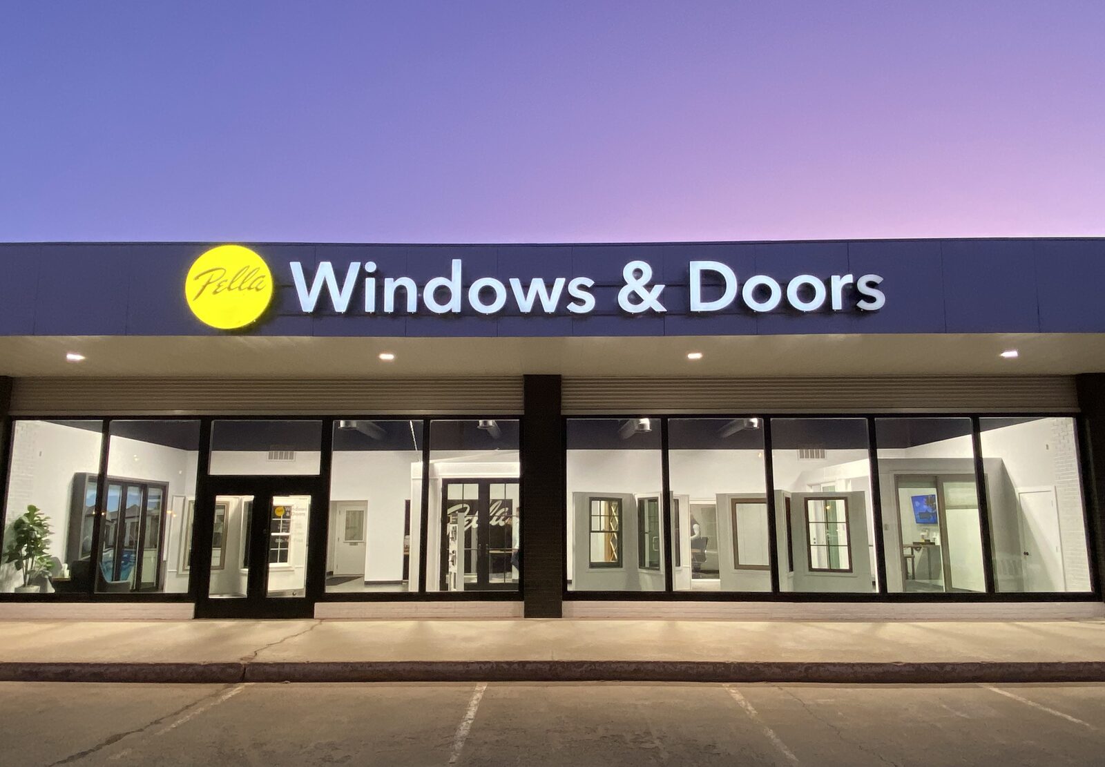 Storefront of Pella Windows & Doors at dusk, showing large glass windows, doors, and illuminated signage on the building.