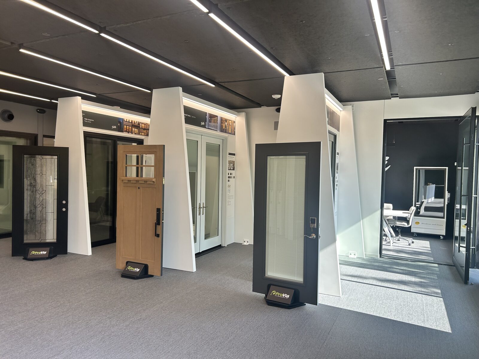 A modern showroom displays various styles of doors on stands, with natural light coming through glass walls and an office area visible in the background.
