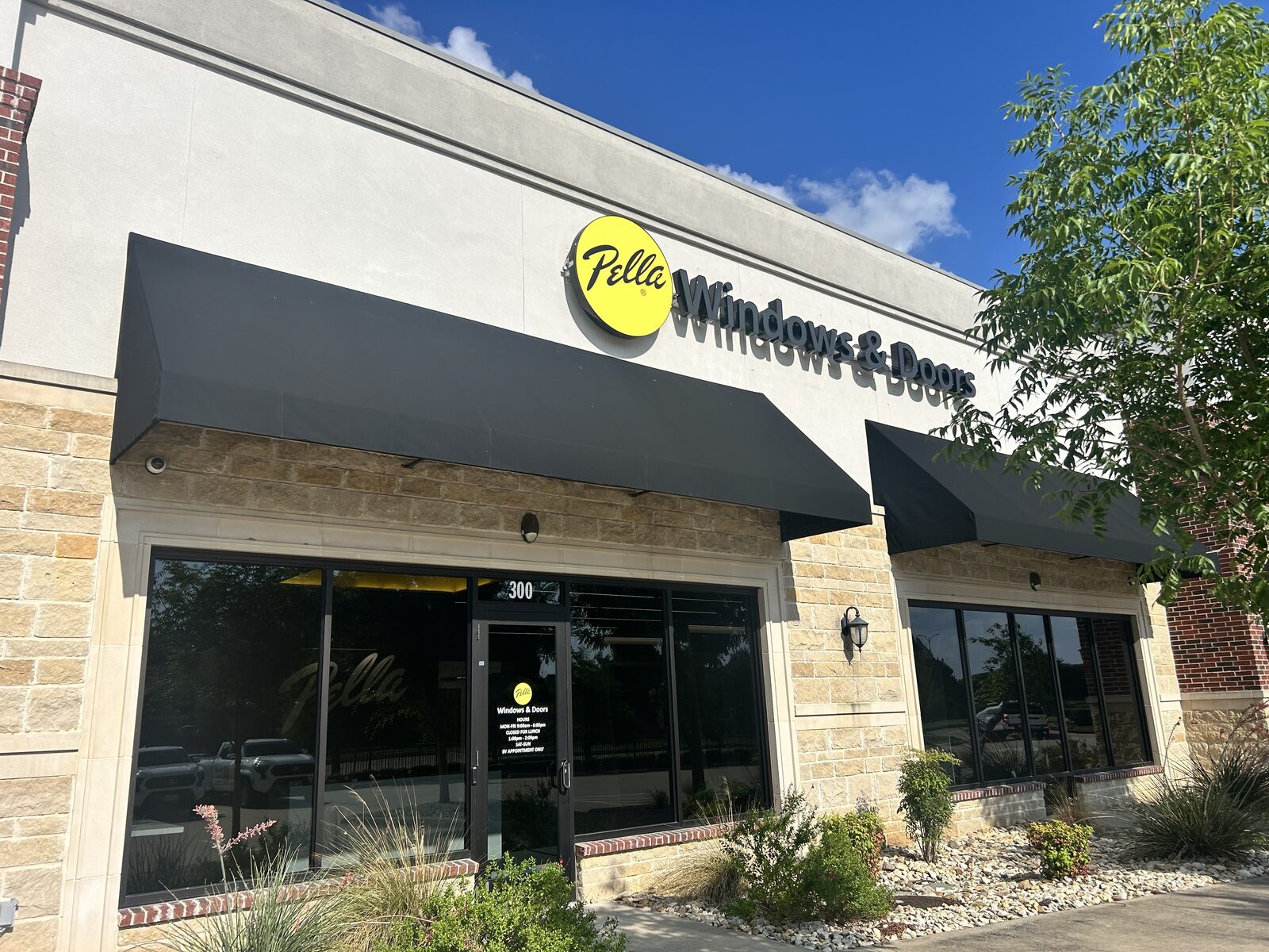 Exterior of a Pella Windows & Doors store with black awnings, large front windows, stone facade, and landscaping under a blue sky.