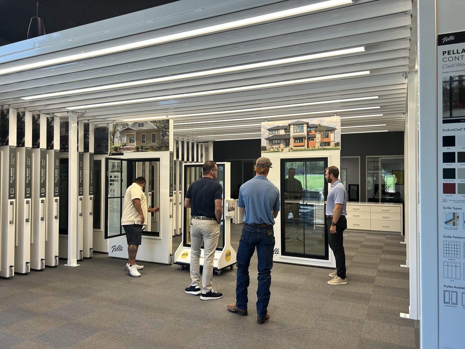 Four men stand in a showroom, looking at window and door displays. The space features sample products, bright lighting, and informational panels on the walls.