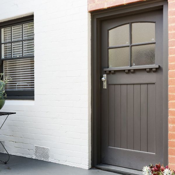 A brown wooden door with glass panes is set in a white brick wall next to a window with closed blinds and a small table with potted plants.