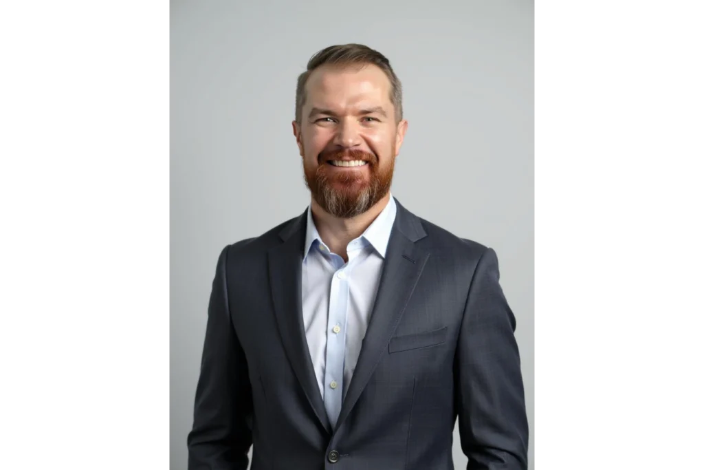 Man with short hair and a beard wearing a gray suit jacket and light blue shirt, standing and smiling against a plain light background.