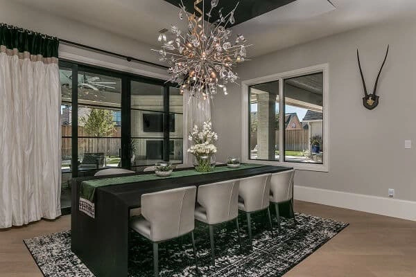 Modern dining room with a dark rectangular table, six light chairs, floral centerpiece, statement chandelier, large windows, and wall-mounted decorative piece above a black-and-white patterned rug.