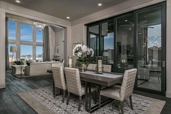 Modern dining room with a table and six chairs, patterned rug, sliding glass doors, and an adjacent living area with large windows and a chandelier.
