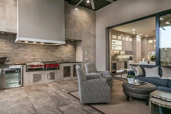 Modern indoor-outdoor living space with built-in kitchen, grill, and seating area; adjacent to a bright, open kitchen.