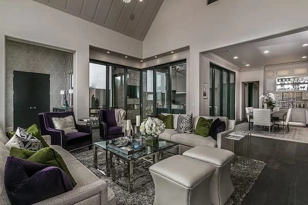Modern open-concept living room with neutral sofas, purple and green accents, glass coffee table, and view into dining area and kitchen. Large glass doors provide natural light.