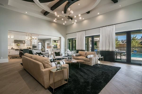 Modern living room with beige sofas, black rug, and large windows; open view to kitchen and outdoor pool area under a high ceiling with exposed beams and a chandelier.